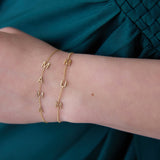 A close-up of a wrist wearing two yellow gold Custom Gold Bracelets in beaded chains, one with four letters M, A, M, A and the other with three letters M, U, M, against a dark turquoise blue silk fabric.
