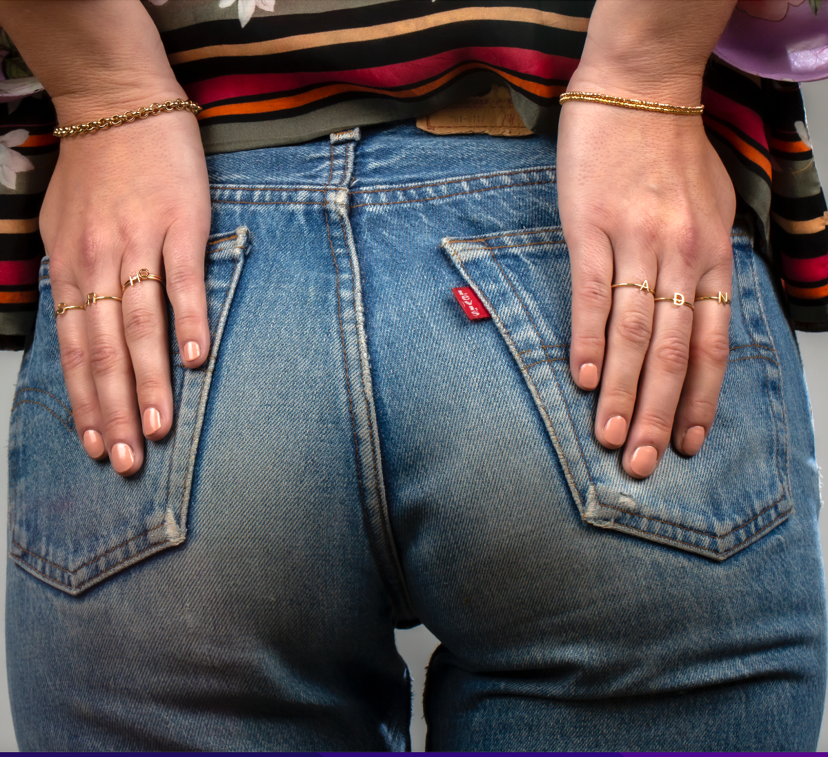 Close-up of a person wearing blue jeans with their hands on the back pockets, wearing gold rings and bracelets including two Diamond Cut Bracelets, a Rolo Bracelet and various Letter Rings.