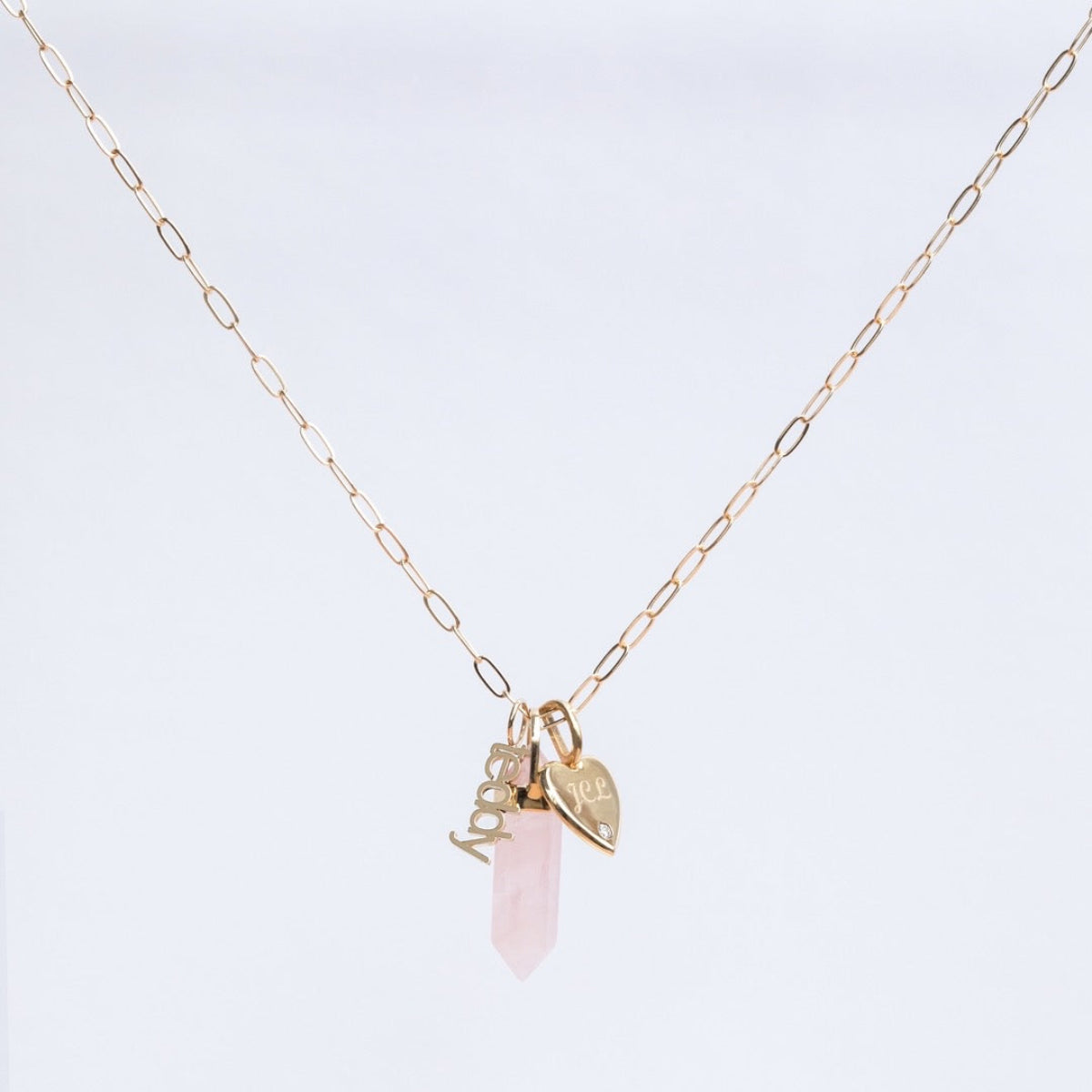 An Element Short Link Chain with a Petite Nameplate Pendant reading “teddy,” an Engraved Heart Charm, and a Balance Crystal Charm, shown against a light grey background.