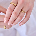 Close-up of hands wearing gold rings, including a mini letter J ring, delicate beaded and twisted bands, a geo stacking ring and a paris ring, on a light neutral background.