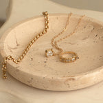 A yellow gold on C'est Moi Link Ring, C'est Moi Deco Necklace and Rolo Necklace in a textured beige bowl. 