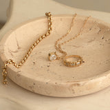 A yellow gold on C'est Moi Link Ring, C'est Moi Deco Necklace and Rolo Necklace in a textured beige bowl. 