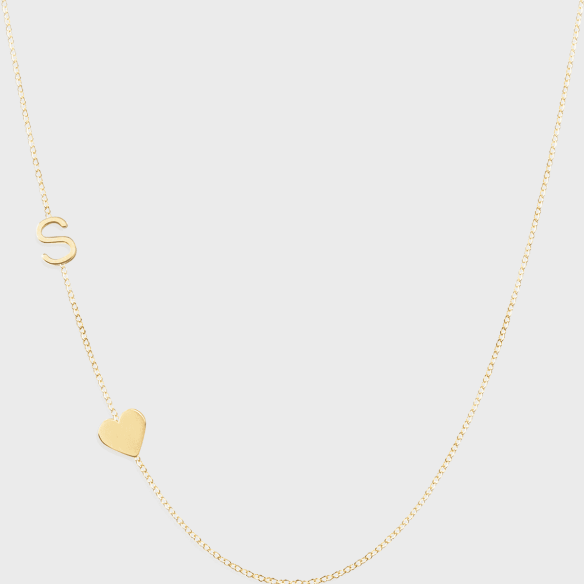 A Monogram Necklace with Heart Yellow Gold and a letter S charm on a light grey background. 