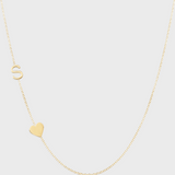 A Monogram Necklace with Heart Yellow Gold and a letter S charm on a light grey background. 