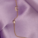 A Monogram Necklace with Diamond and the letter E in yellow gold, against light purple silk fabric as the background.