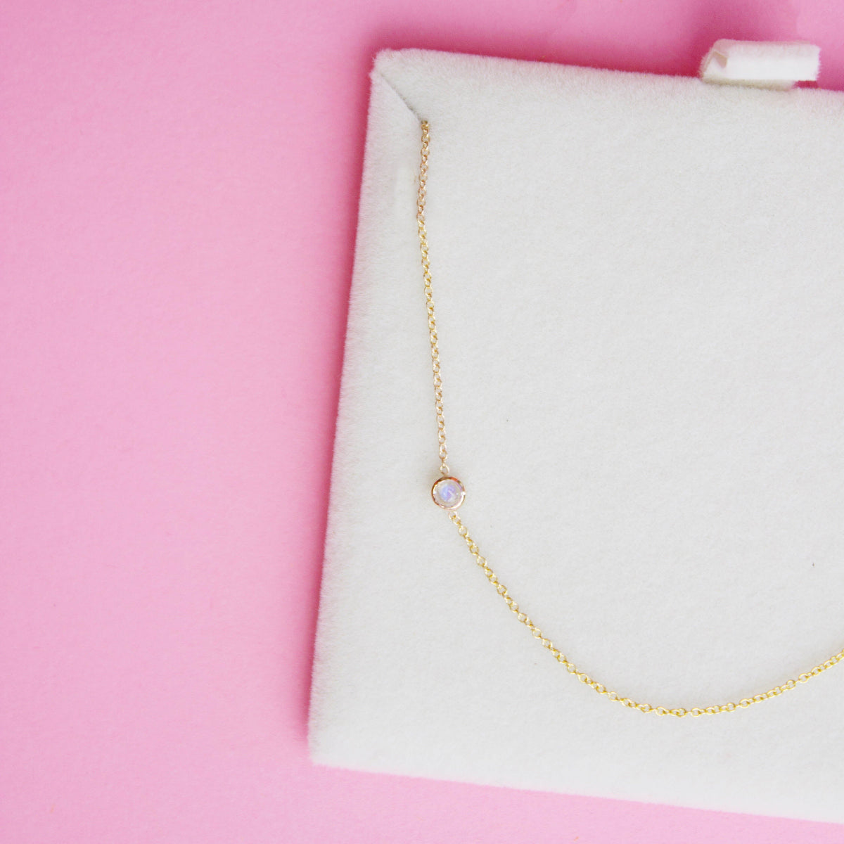 A 14K Gold Asymmetrical Birthstone Necklace - Moonstone (June) in a white cushion case, against a pink background. 