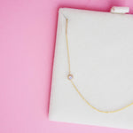 A 14K Gold Asymmetrical Birthstone Necklace - Moonstone (June) in a white cushion case, against a pink background. 
