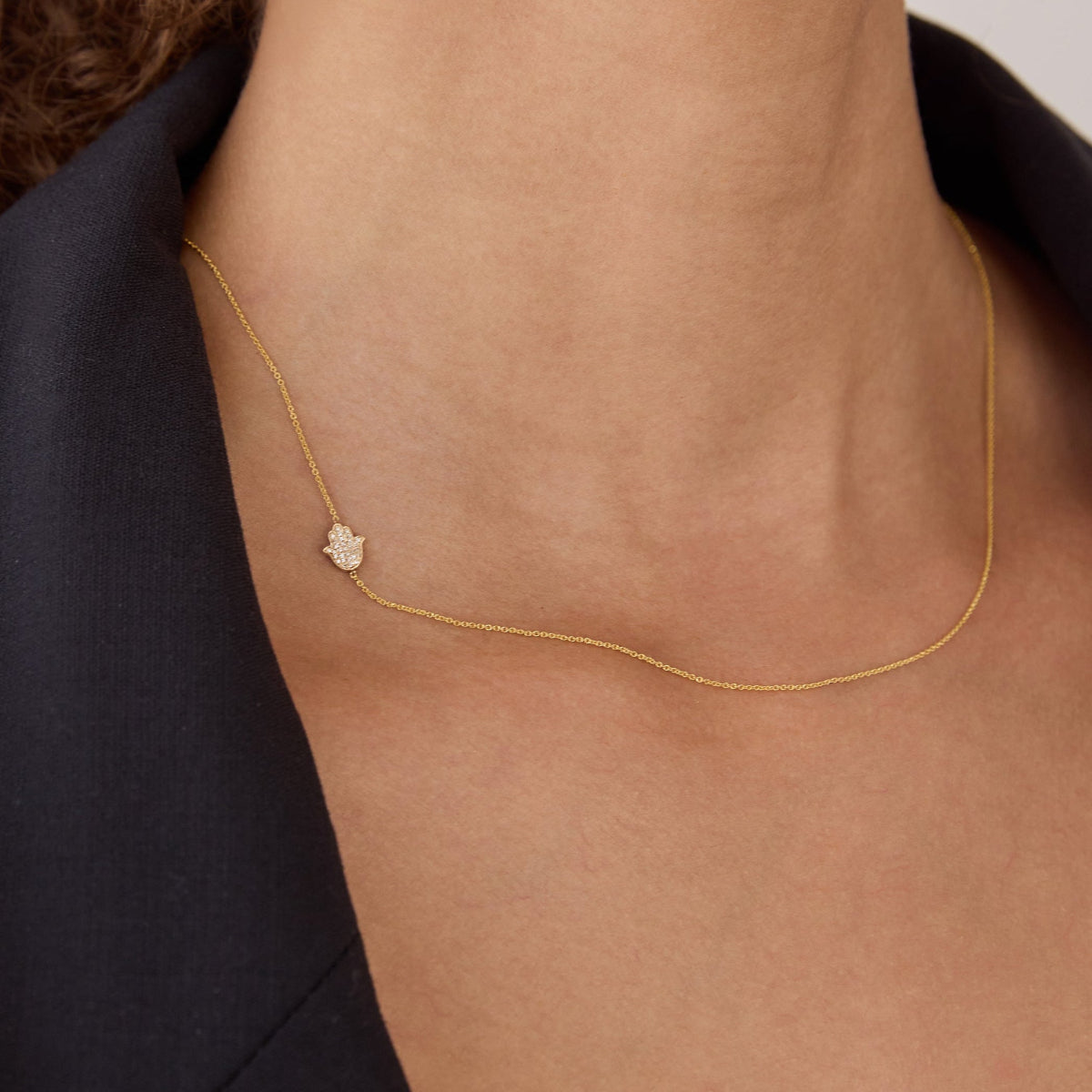 A close-up of a woman's neck wearing a yellow gold cable chain 14k Gold Asymmetrical Charm Necklace - Pavé Hamsa, styled with a black jacket. 