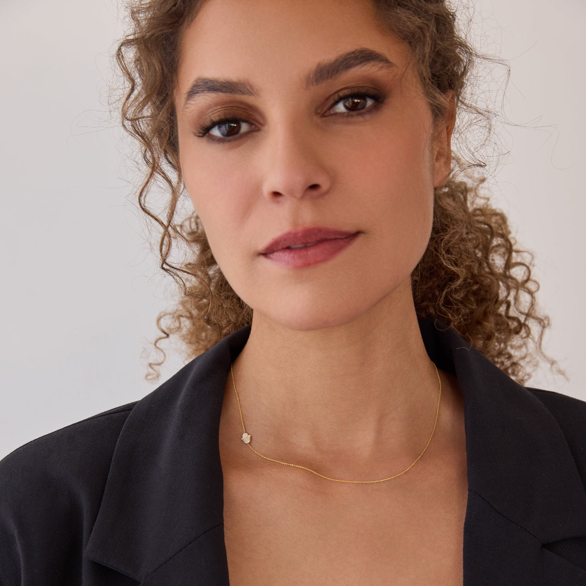 A woman wearing a yellow gold 14k Gold Asymmetrical Charm Necklace - Pavé Hamsa with a cable chain styled  which a black blazer against a white background. 