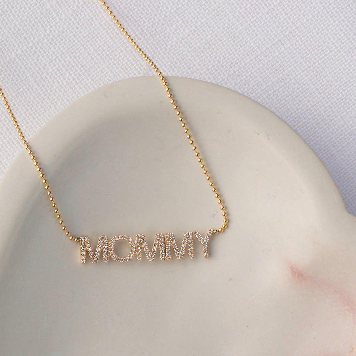 A Pavé MOMMY Necklace in yellow gold on a cable chain laid on top of a cream surface and white fabric background. 