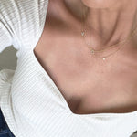 Close-up of a person wearing an off-white top and three yellow gold necklaces – one with a letter V charm, one with a letter O charm, and one with a birthstone.