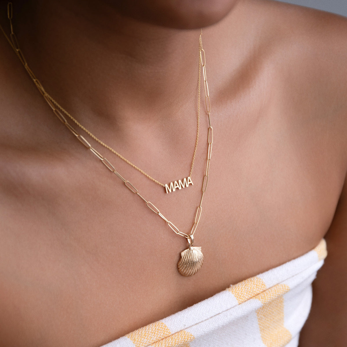 A close-up of a woman's décolletage wearing a MAMA Necklace and an Element Long Link Chain with a Gold Scallop Shell, with a white and light orange strapless top. 