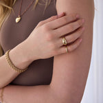 Close-up of a hand wearing a yellow gold Paris Ring, a Tube Ring and two Rolo Bracelets, styled with a brown top and the Peony Bouquet Necklace with a Diamond Heart Locket Charm, against a white background.
