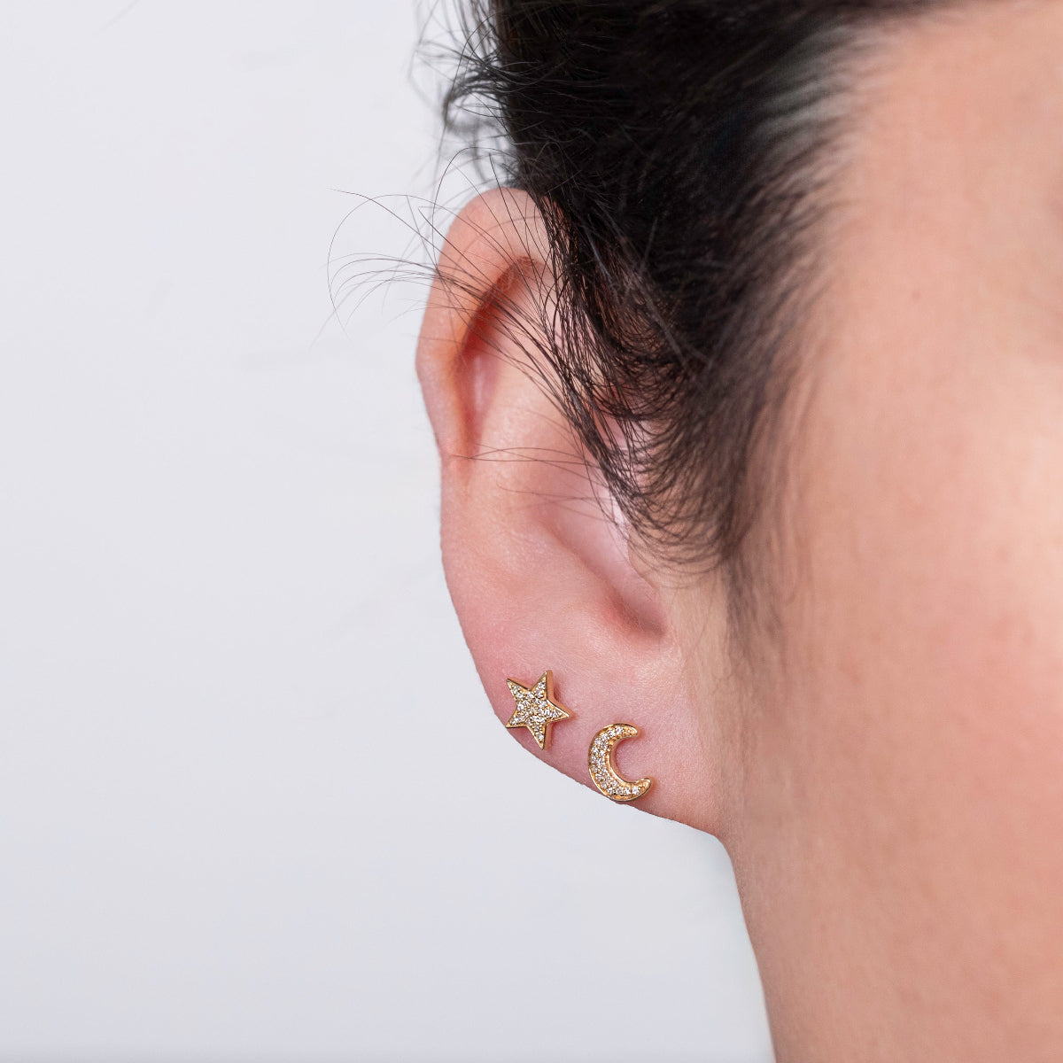 Close-up of yellow gold Pave Charm Studs - Moon and Star in a woman's ear against a light grey background. 