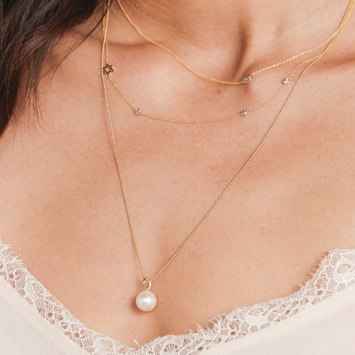 Close-up of a woman's decellotage wearing a 14K Gold Asymmetrical Charm Necklace -Angel Wing, a Custom Gold Necklace - 4 Letter,  and a Perla Pendant on a Classic Bead Chain, with a white lace top. 