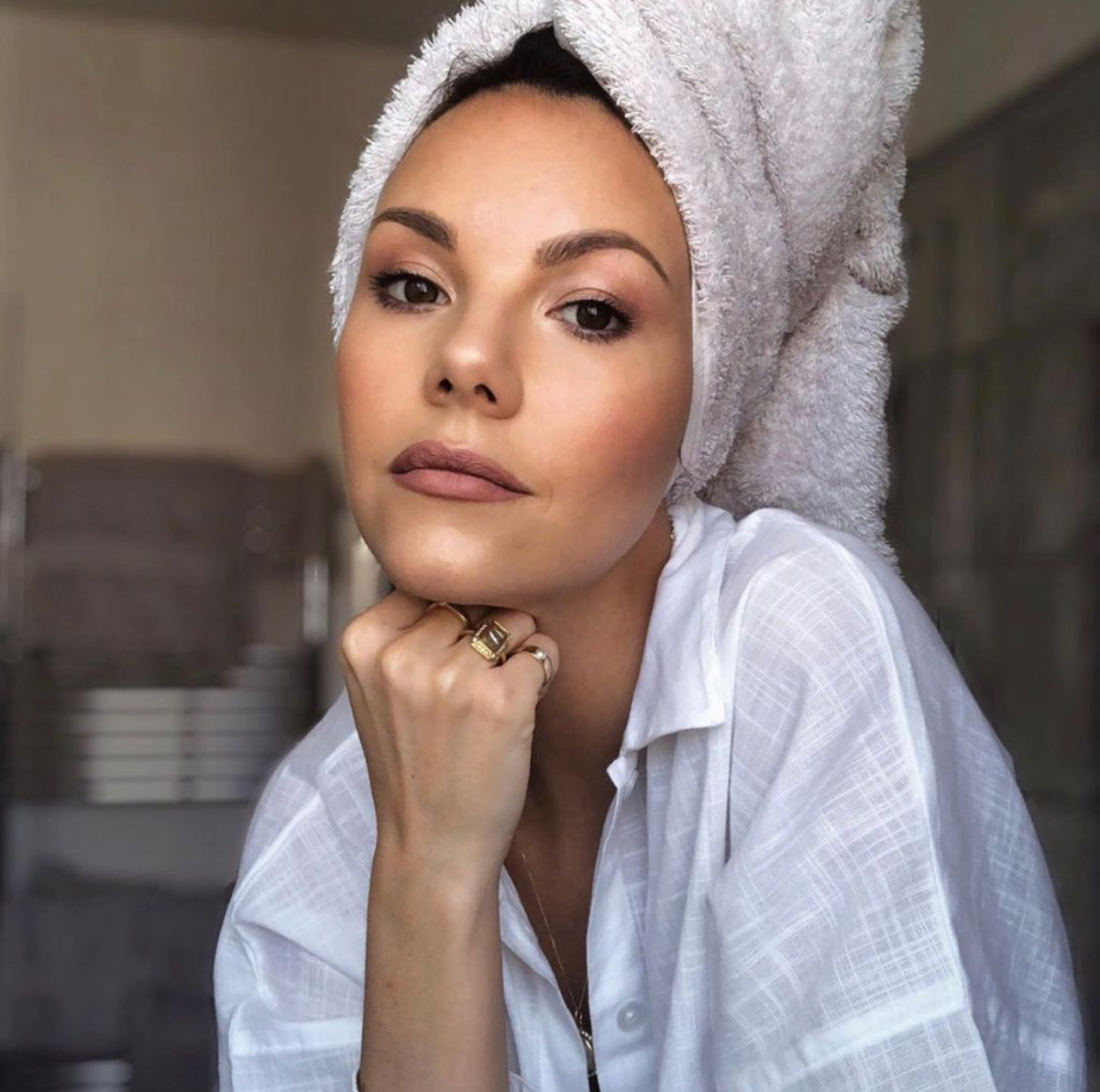 A woman wearing several rings, including a Perla Pinky Ring with a towel  wrapped around her head, wearing a white shirt in a blurred indoor setting. 