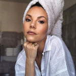 A woman wearing several rings, including a Perla Pinky Ring with a towel  wrapped around her head, wearing a white shirt in a blurred indoor setting. 