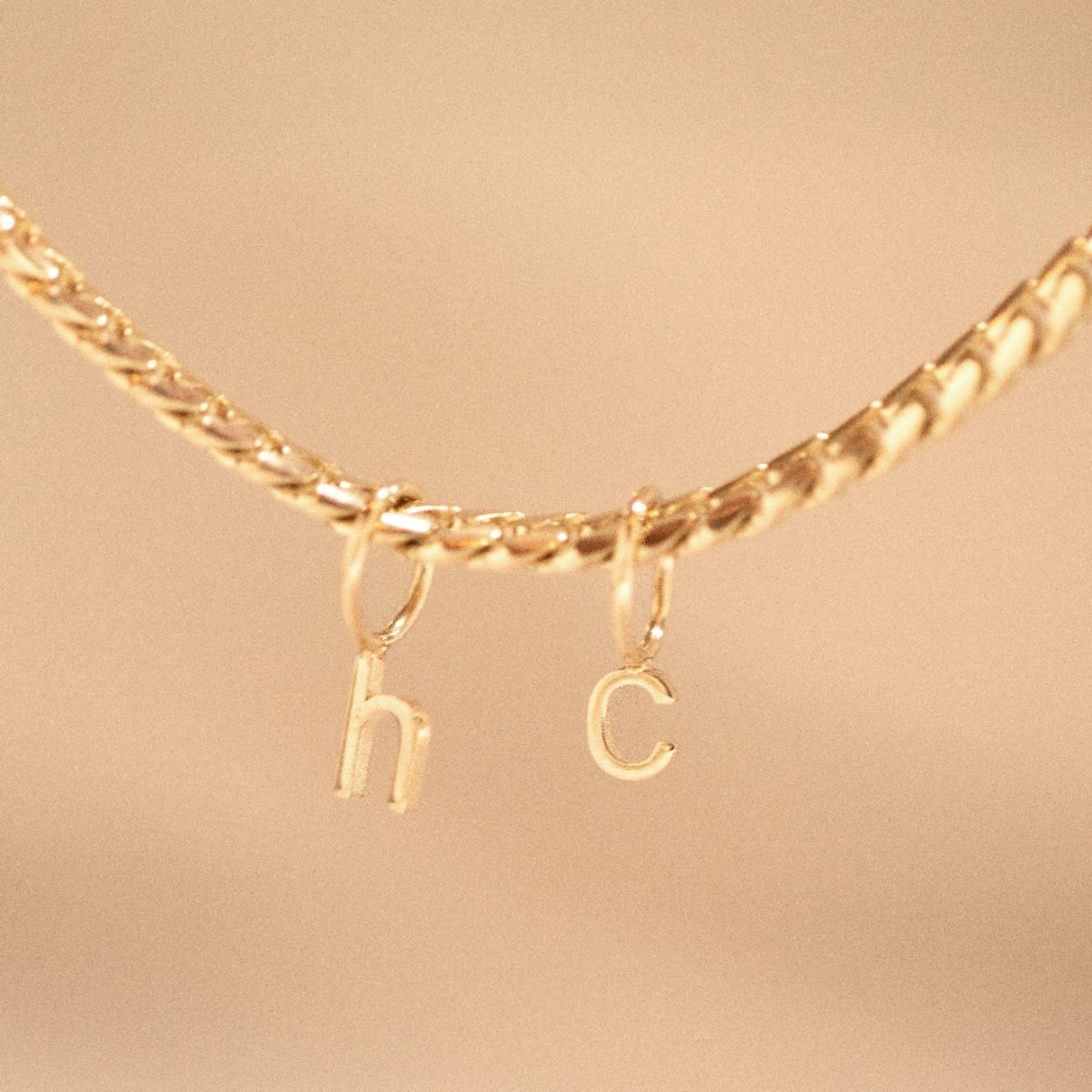 A yellow gold necklace with Petite Letter Charms in h and c on against a beige background.