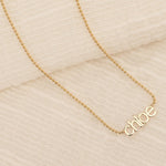 A yellow gold Petite Nameplate Necklace with 'chloe' against a cream fabric background. 