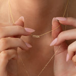 A close-up of a neck wearing a yellow gold Petite Nameplate Necklace with “max,” held by hands, styled with another longer yellow gold necklace.  
