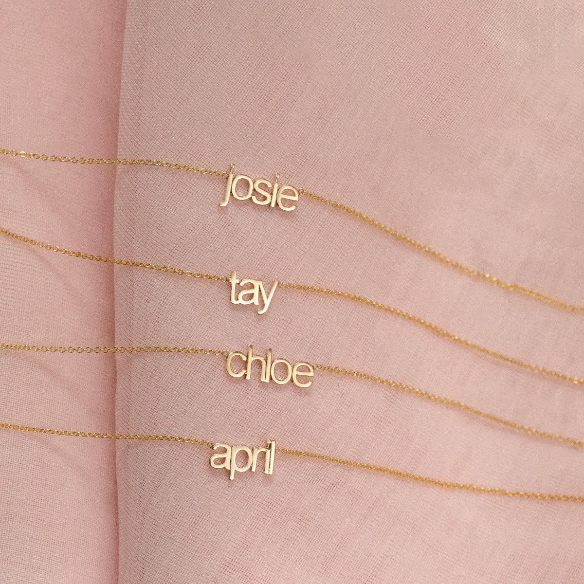 Four Petite Nameplate Necklaces in yellow gold, in the names Josie, Tay, Chloe and April (all in lowercase) on a pink fabric background. 