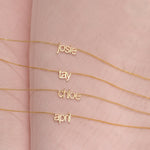 Four Petite Nameplate Necklaces in yellow gold, in the names Josie, Tay, Chloe and April (all in lowercase) on a pink fabric background. 
