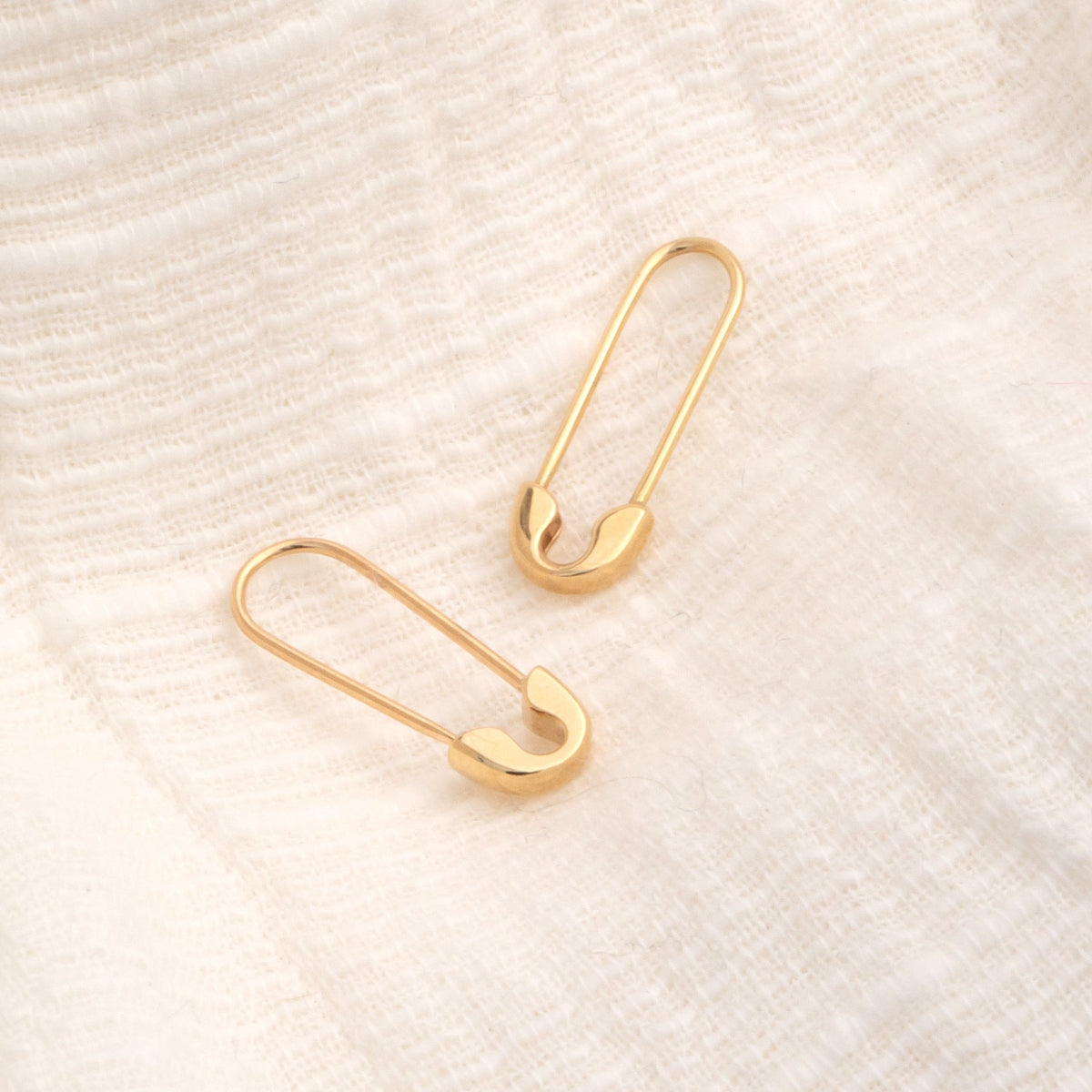Two Gold Safety Pin Earrings on a textured cream background. 