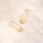 Two Gold Safety Pin Earrings on a textured cream background. 