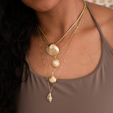 A close-up of a woman wearing a Chic Cuban Curb Chain with The Golden Venus Shell, an Element Long Link Chain with a Golden Scallop Shell, a Classic Bead Chain with a Golden Coast Shell, and The Slide Chain with a Golden Spiral Shell, with a light brown singlet top. 
