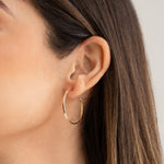 Close-up of woman's ear wearing Sleek & Chic Hoops - Small in yellow gold with her brown hair visible against a light grey background. 
