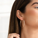 Close-up of a woman's ear wearing a Sleek & Chic Hoops - Medium in yellow gold, with her brown hair and a white background. 