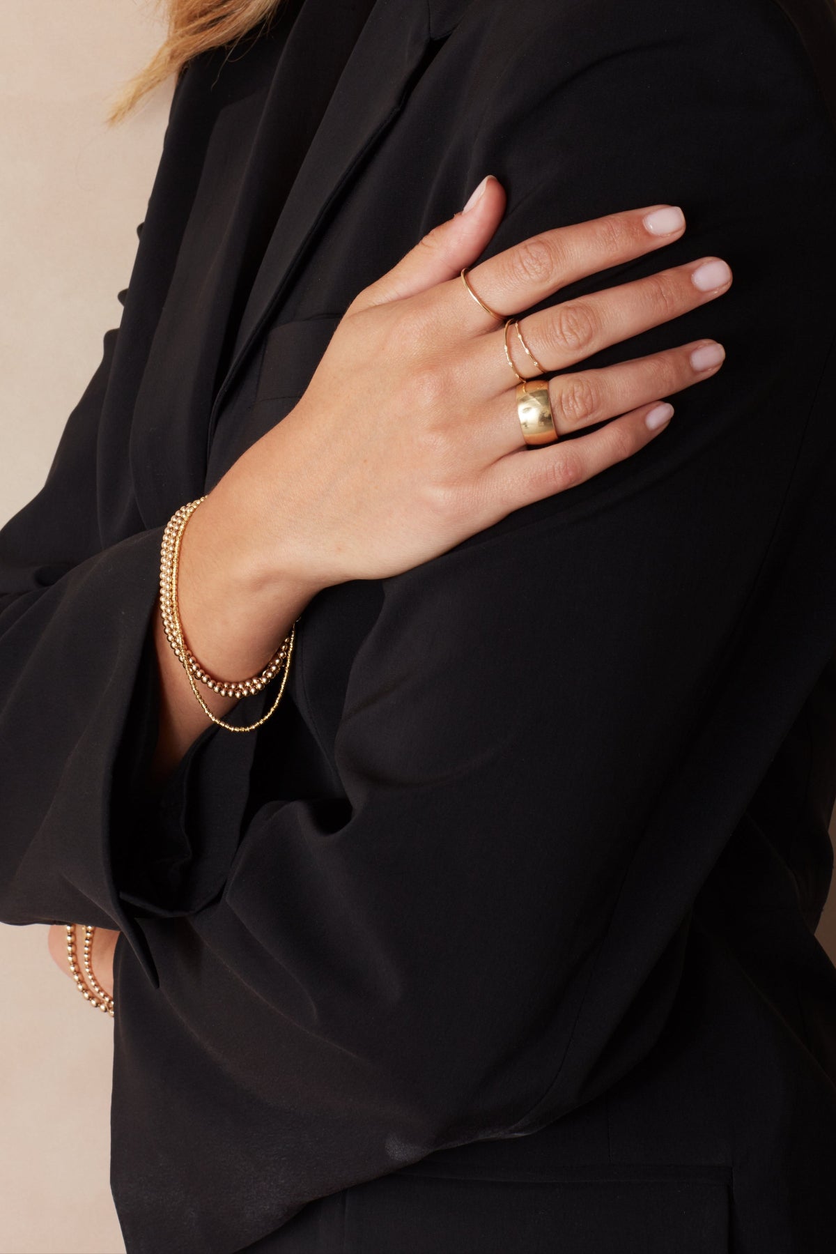 A woman's hand wearing two 4 Diamond Rings, a Thick Band and Delicate Band, with Bubble Bracelets and a Diamond Cut Bracelet, all in yellow gold, resting on her black blazer against a light grey background.
