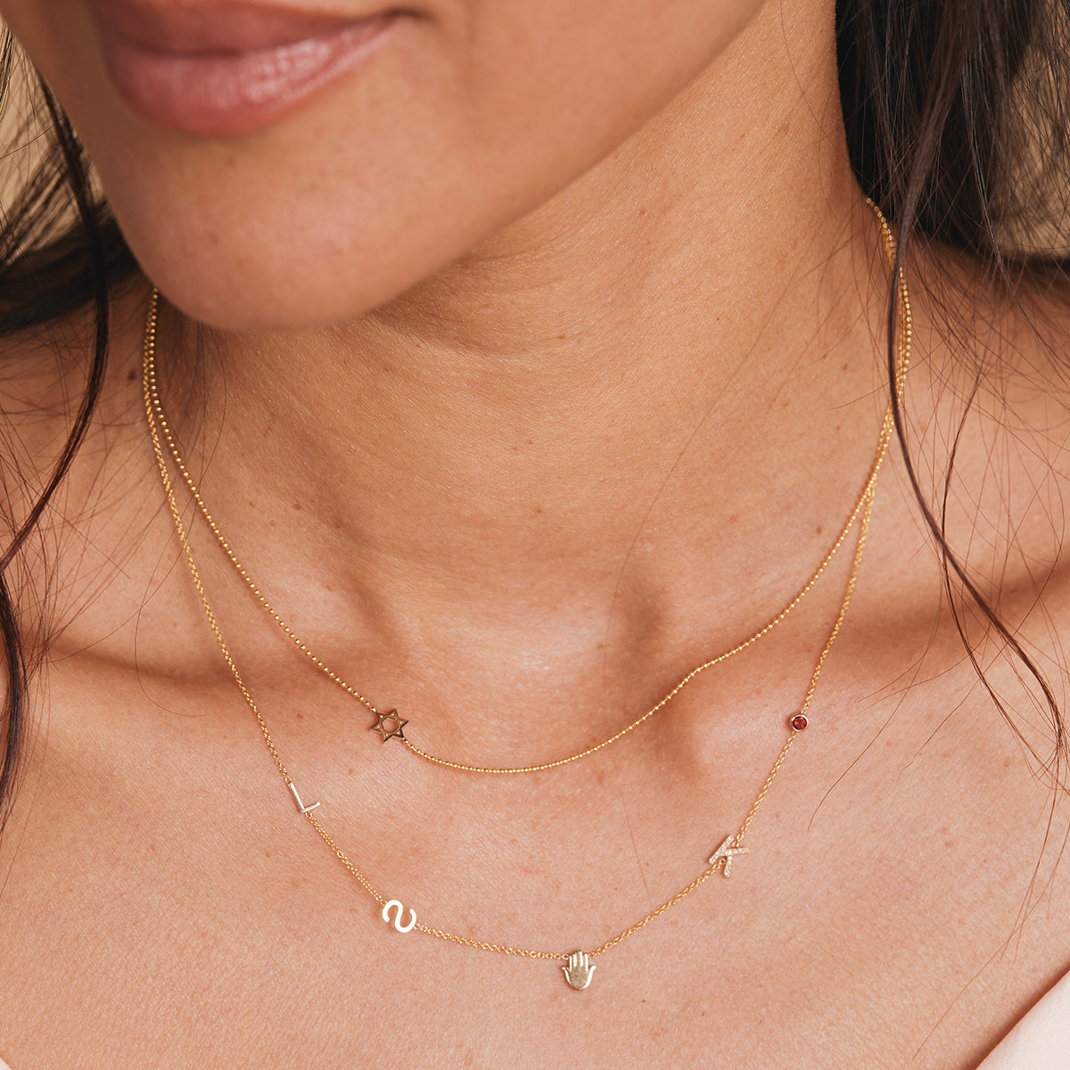 A close-up of a woman’s neck wearing a Custom Gold Necklace with five charms (letters L, S, and K, a hand and a birthstone) layered with a Charm Necklace featuring a Star of David charm, both in yellow gold.