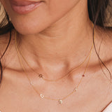 A close-up of a woman’s neck wearing a Custom Gold Necklace with five charms (letters L, S, and K, a hand and a birthstone) layered with a Charm Necklace featuring a Star of David charm, both in yellow gold.