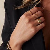A woman’s hand wearing two 4 Diamond Rings, a Thick Band, a Delicate Band, Bubble Bracelets, a Diamond Cut Bracelet, and an Element Long Link Chain, all in yellow gold, styled with a black blazer against a light grey background.