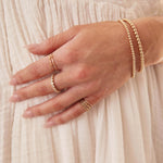 A woman’s hand resting on her cream dress, wearing gold jewellery including Bubble Bracelets, an R Letter Ring, a Number 1 Ring, Delicate Trio Stacking Rings, and a Magnolia Bouquet Ring.