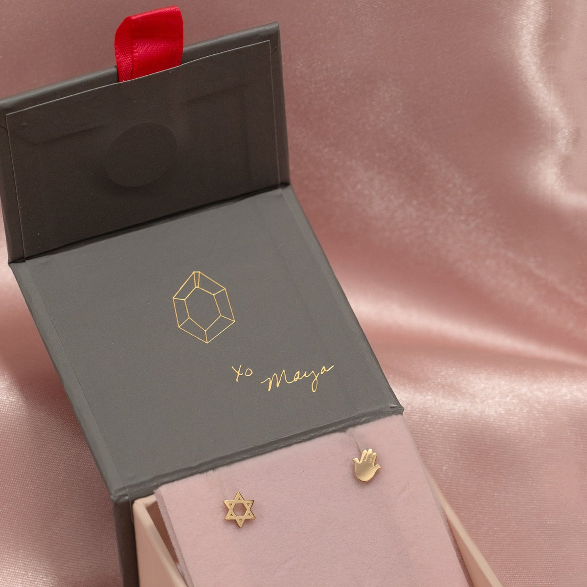 Charm Studs - Star of David and Hamsa in yellow gold in a Maya Brenner jewellery box against a light pink silk background. 