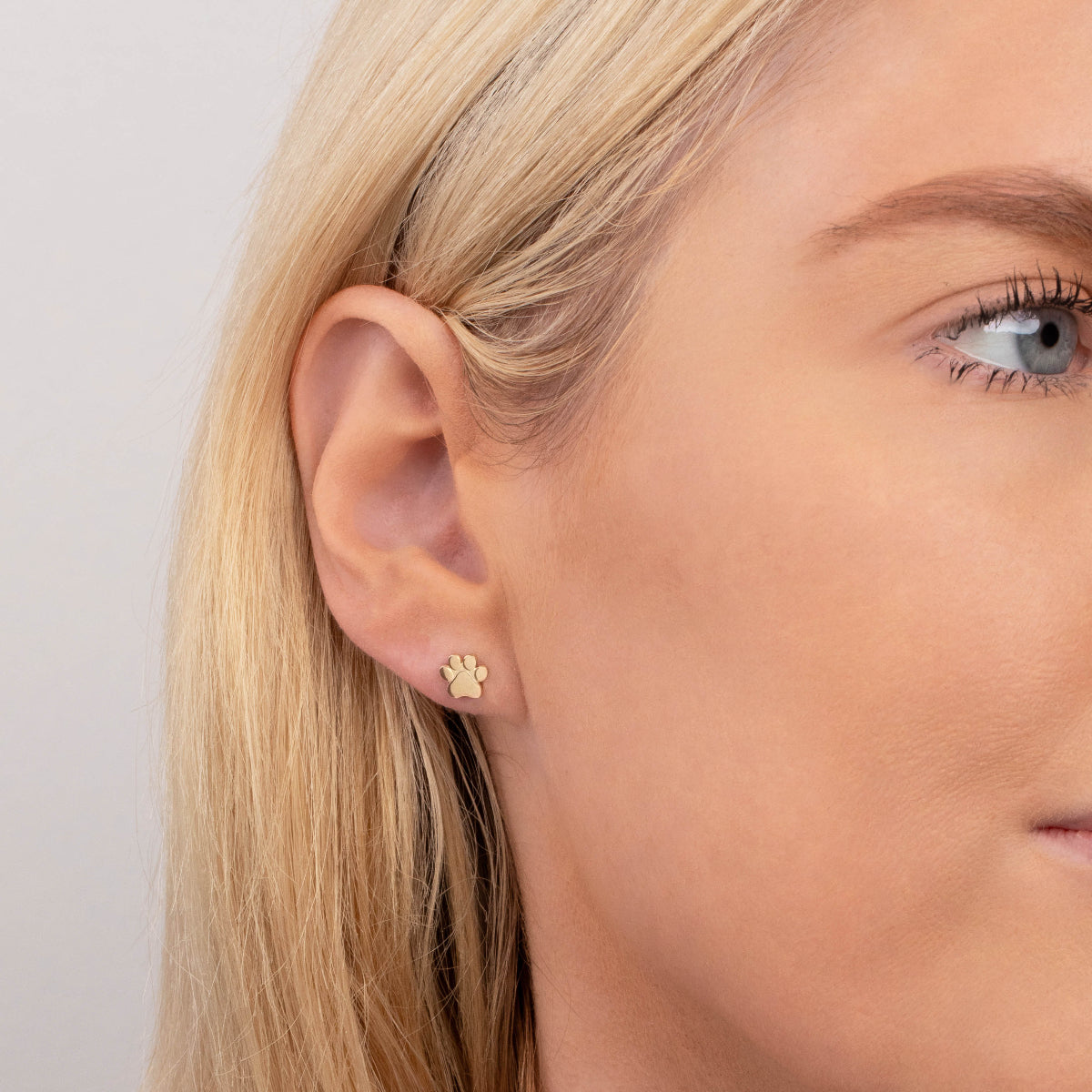 Close-up of a yellow gold Charm Stud - Paw in a blonde woman's ear with a light grey background. 