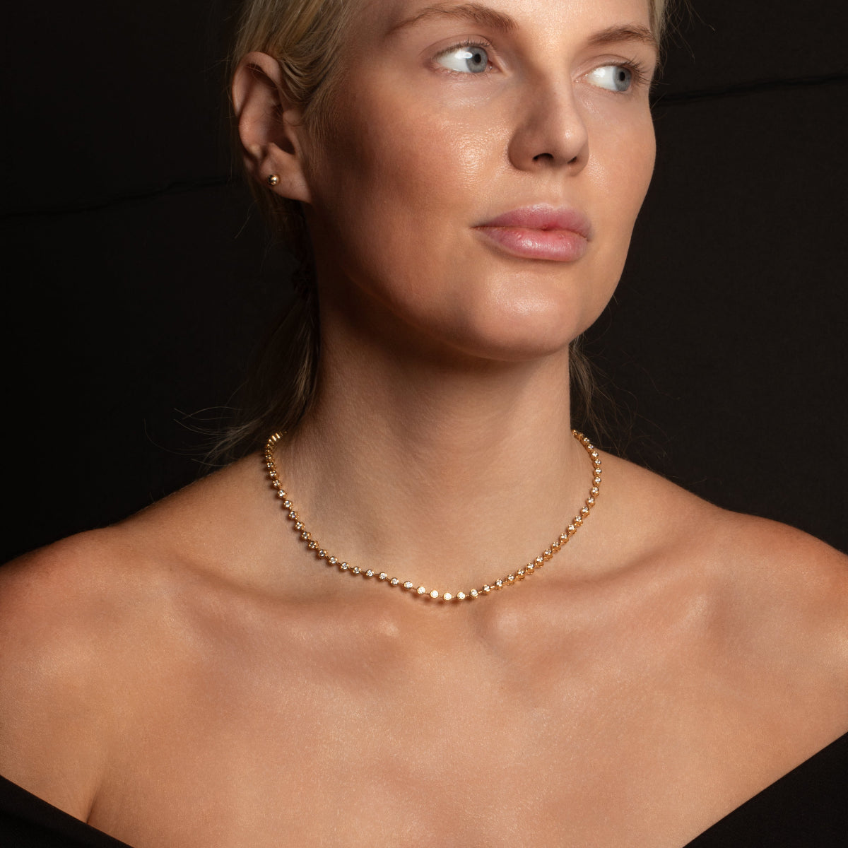 A blonde woman wearing The Ultimate Diamond Necklace in yellow gold with a black background. 