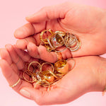 A pair of hands holding several yellow gold and white gold rings, including Thick Bands, Delicate Trio Stacking Rings, Braided Rings, Flat Rings, against a light pink background. 