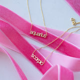 Petite Zodiac Necklaces 'aquarius' and 'scorpio' in yellow gold displayed on top of pink ribbon against a light grey background. 