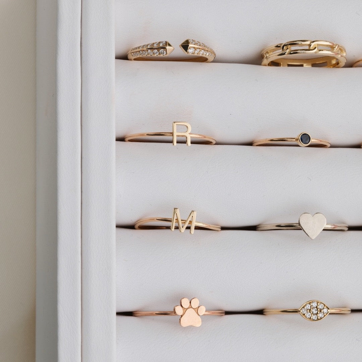 A number of yellow gold and rose gold rings in a white case, including an Open Diamond Ring, a Link Ring, an R Letter Ring, a Birthstone Ring, an M Letter Ring, a Heartfelt Ring, a paw ring, and a Pavé Petal Ring.