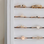 A number of yellow gold and rose gold rings in a white case, including an Open Diamond Ring, a Link Ring, an R Letter Ring, a Birthstone Ring, an M Letter Ring, a Heartfelt Ring, a paw ring, and a Pavé Petal Ring.