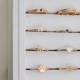 A number of yellow gold and rose gold rings in a white case, including an Open Diamond Ring, a Link Ring, an R Letter Ring, a Birthstone Ring, an M Letter Ring, a Heartfelt Ring, a paw ring, and a Pavé Petal Ring.