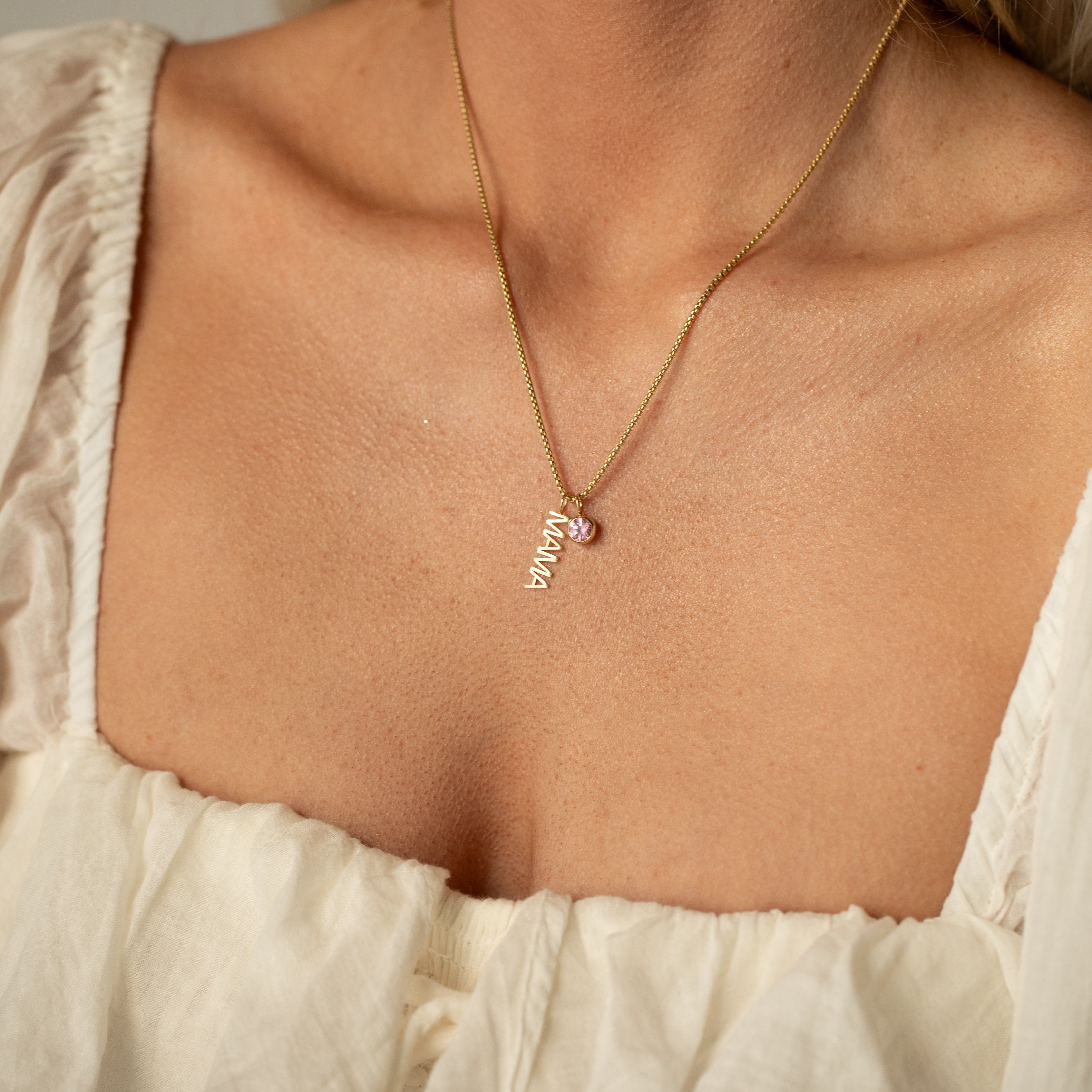 Gold necklace with a 14k gold 'mama' pendant styled with a light pink gemstone pendant on a person wearing a light-colored top.