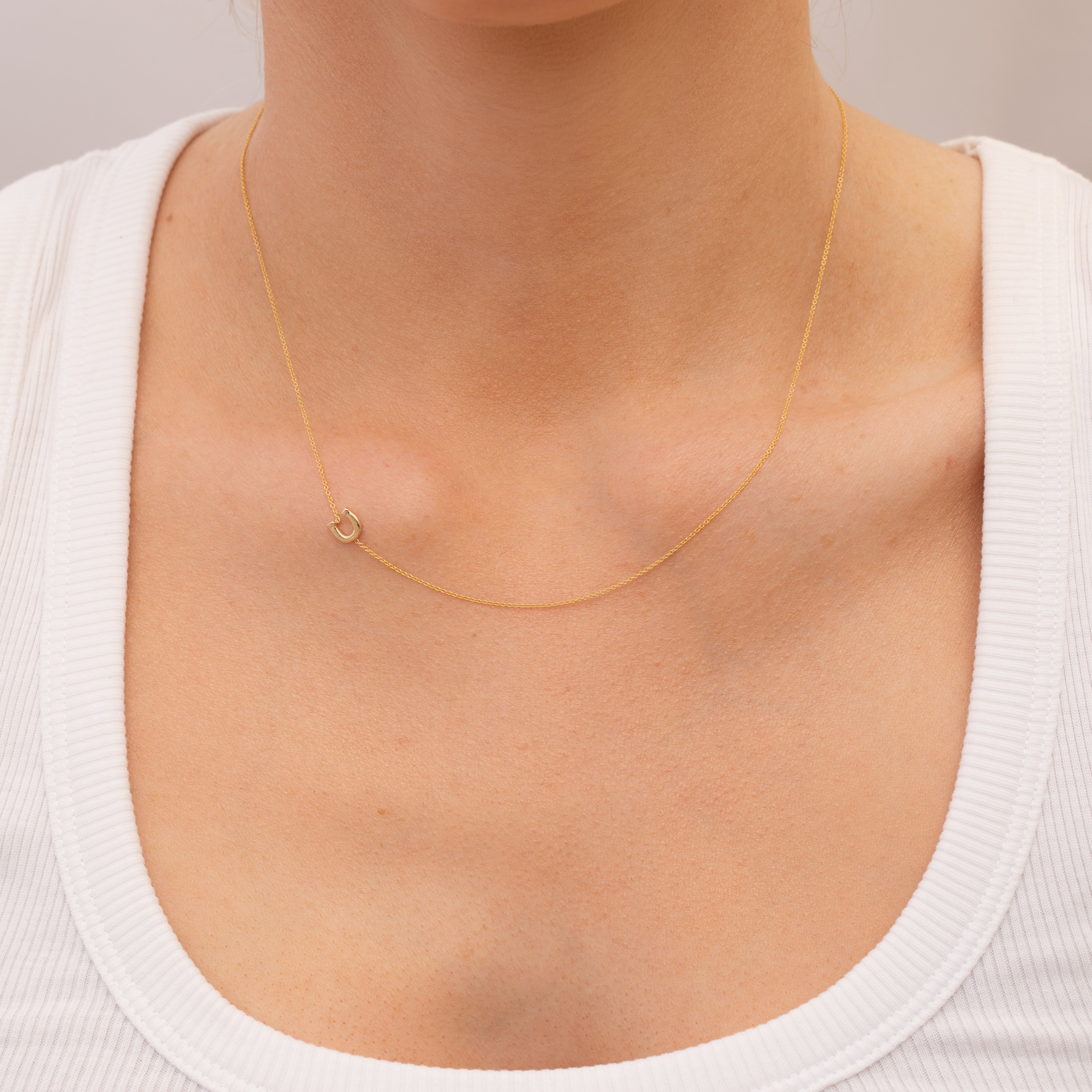 Gold necklace with a small horseshoe charm on a person wearing a white top against a neutral background