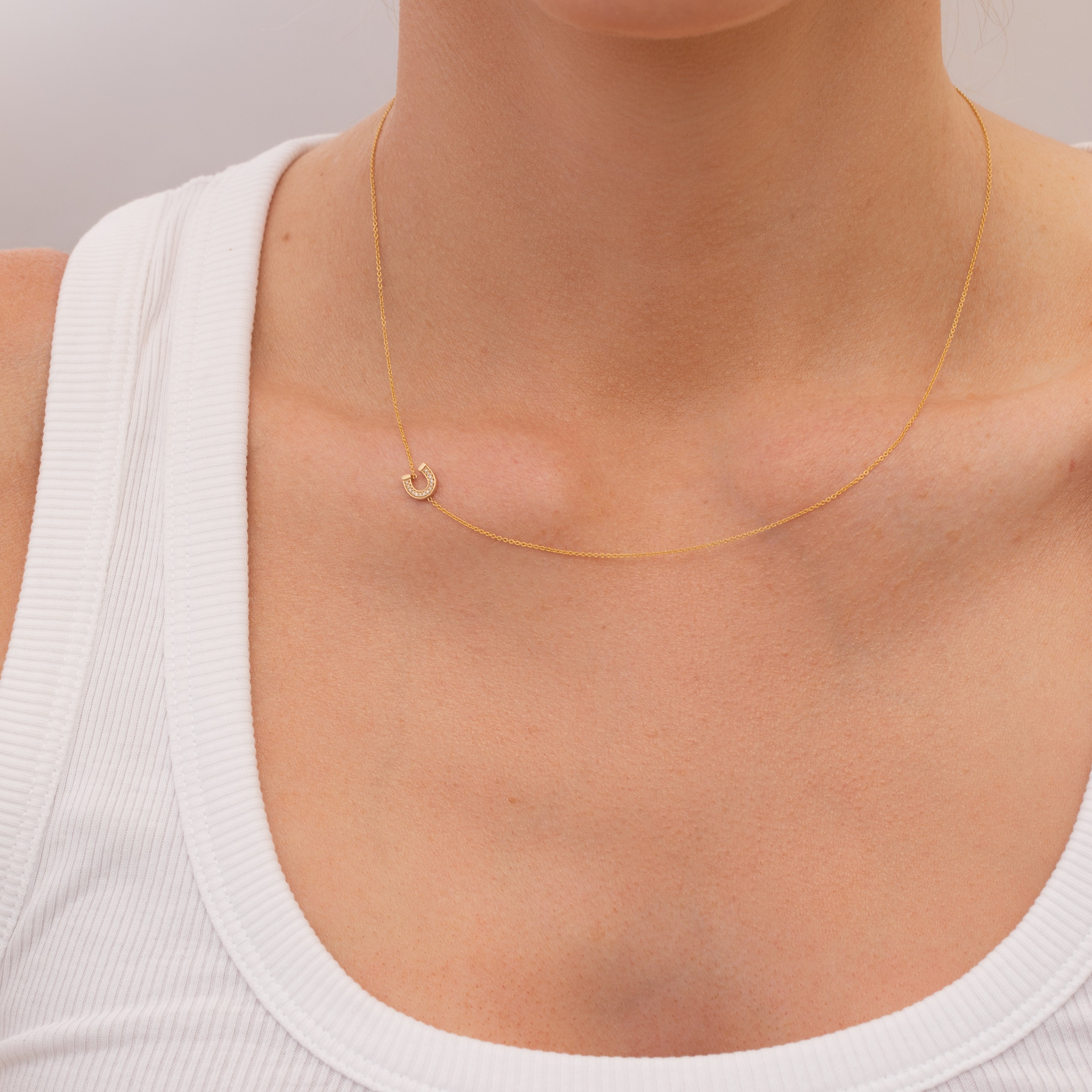 Gold necklace with a small pavé diamond horseshoe charm  on a person wearing a white top against a neutral background
