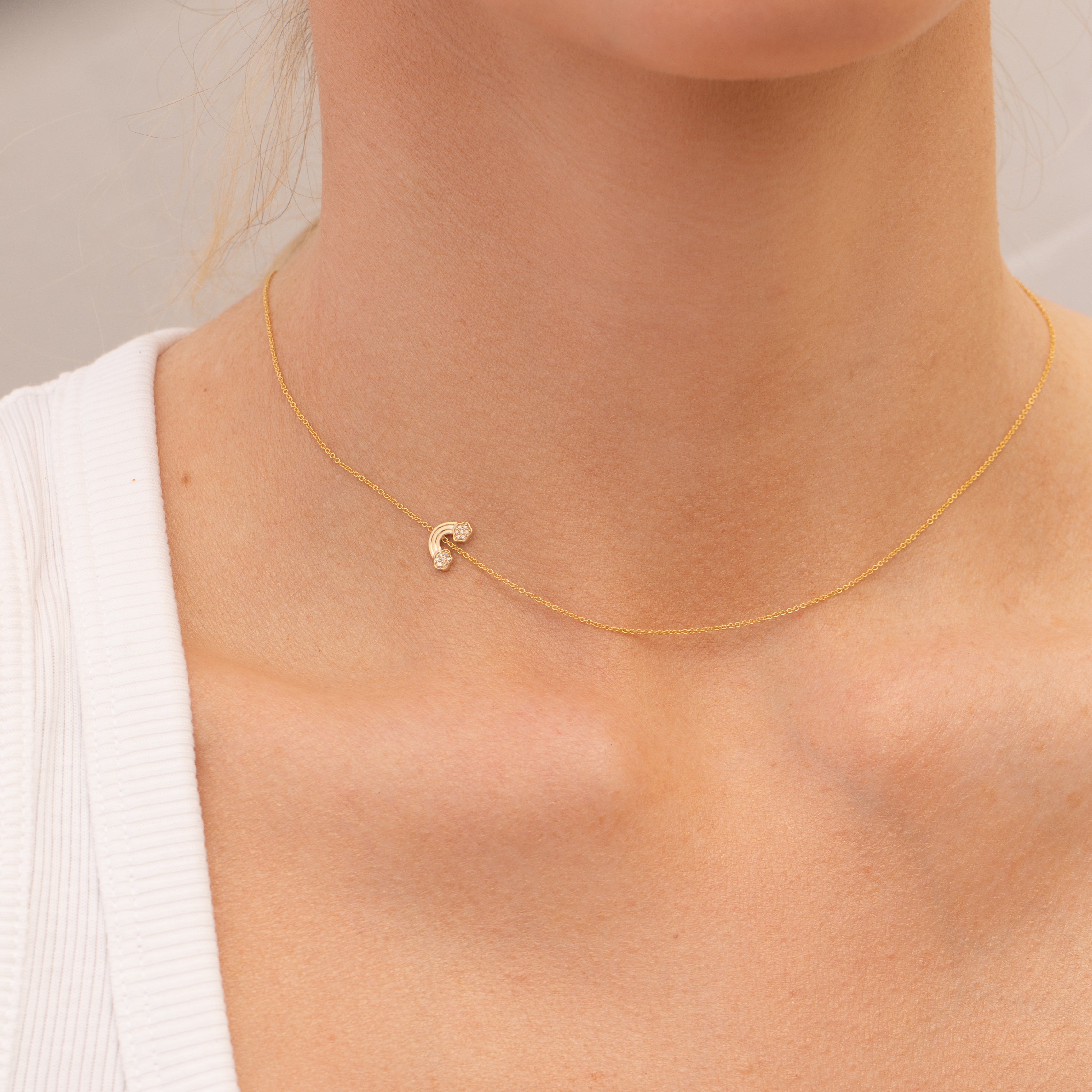 Gold necklace with a small pavé diamond rainbow charm on a person wearing a white top.