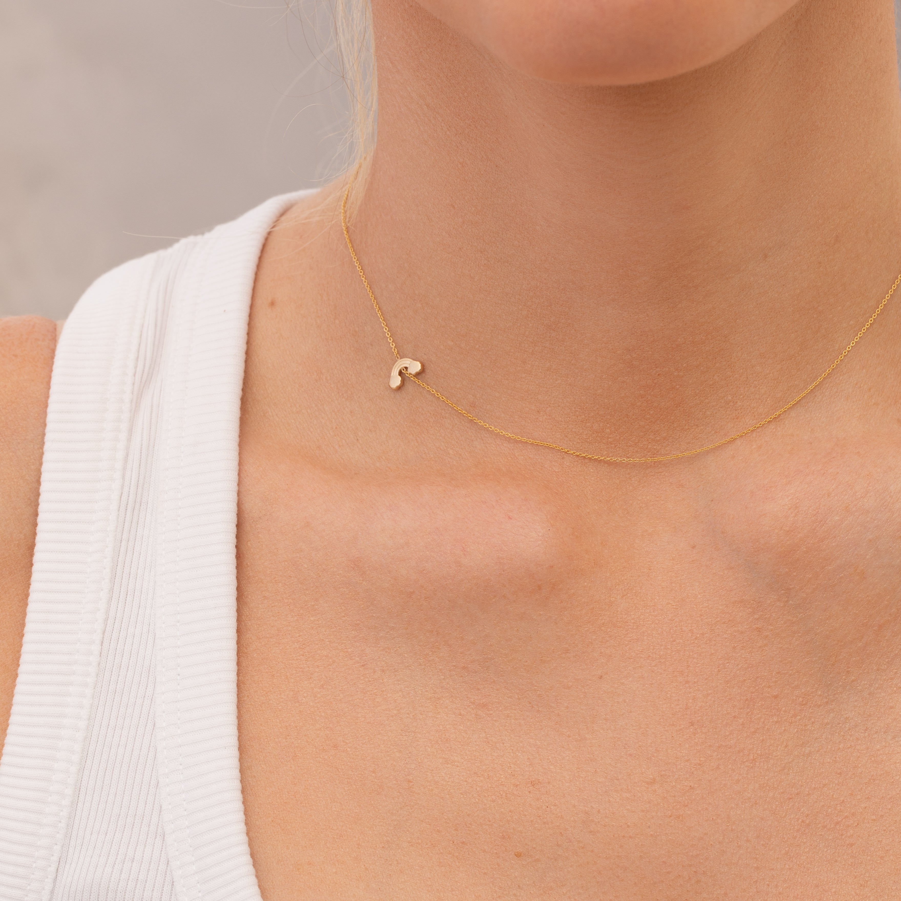 Gold necklace with a small rainbow charm  worn by a person against a neutral background
