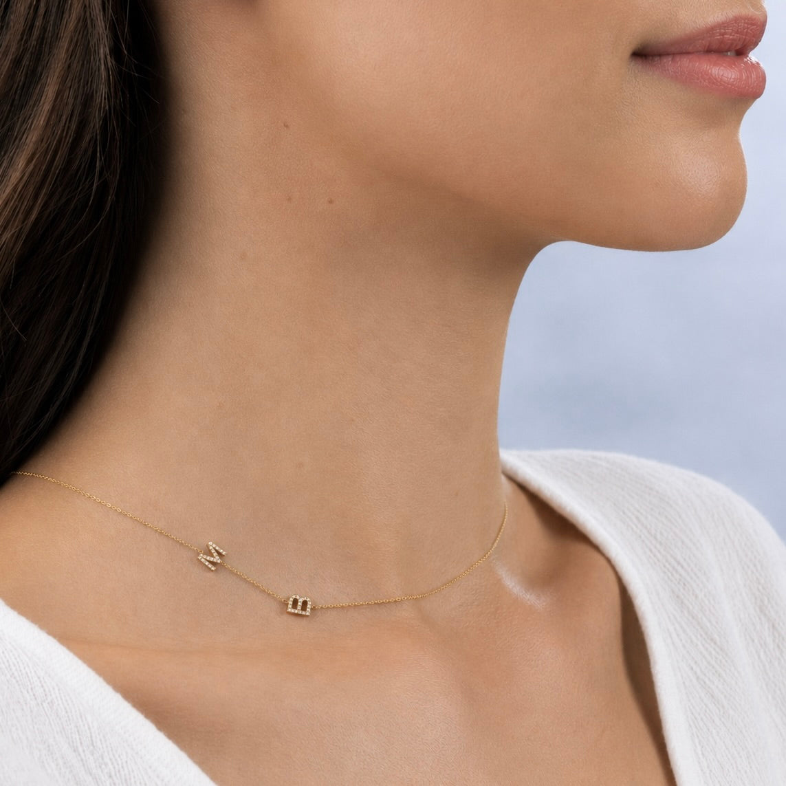 Gold necklace with small letter pendants on a person wearing a white top against a light blue background